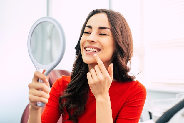 Una chica guapa con un suéter escarlata sostiene un espejo en su mano elegante y mira a través del resultado perfecto del trabajo dental.