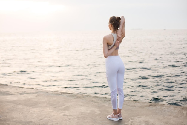 Chica guapa en top deportivo y polainas blancas cogidos de la mano detrás de la espalda con vista al mar en el fondo. Mujer joven practicando yoga junto al mar