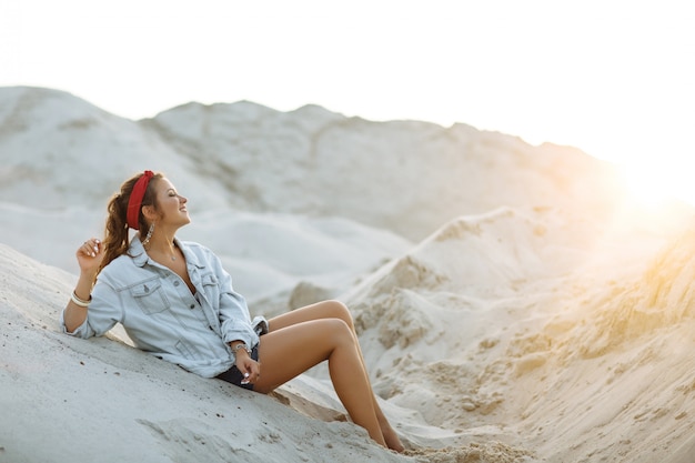 Una chica guapa con vaqueros, pantalones cortos, un aro rojo y un sostén blanco está sentada en la arena.