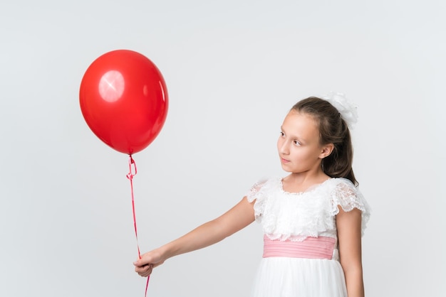 Chica guapa con vestido blanco sosteniendo un globo rojo en la mano extendida mirando cuidadosamente el globo