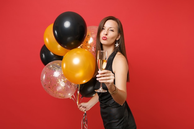 Chica guapa en vestido negro celebrando los labios que soplan enviar beso de aire sostenga una copa de champán globos de aire aislados sobre fondo rojo. Concepto de fiesta de vacaciones de maqueta de cumpleaños de feliz año nuevo de San Valentín.