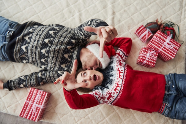 La chica se ha preguntado cara como esta para la selfie imaginaria. Vista superior de la pareja en ropa de Navidad yace en el suelo con regalos.