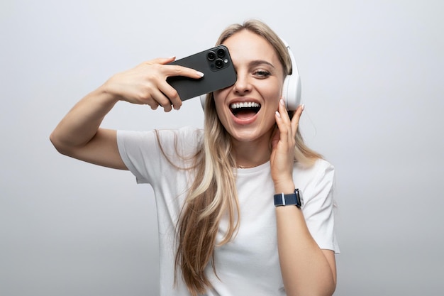 Chica hace una cara en auriculares inalámbricos con un teléfono inteligente en sus manos sobre un fondo blanco.