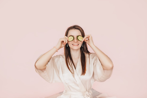 Chica hacer máscaras de belleza facial caseras. Pepinos por el frescor de la piel del contorno de ojos. La mujer cuida la piel joven. Modelo riendo y divirtiéndose en el spa sobre fondo rosa