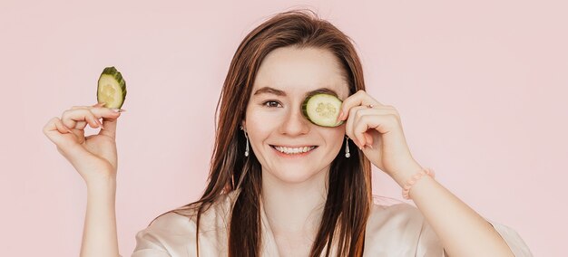 Chica hacer máscaras de belleza facial caseras. Pepinos por el frescor de la piel del contorno de ojos. La mujer cuida la piel joven. Modelo riendo y divirtiéndose en el spa sobre fondo rosa