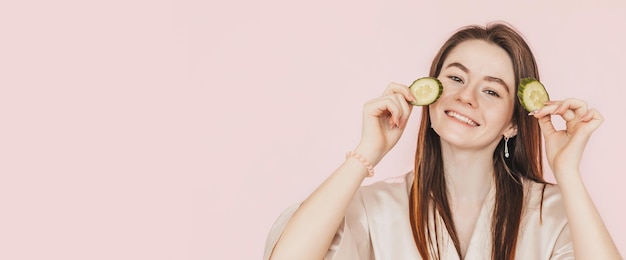 Chica hacer mascarillas caseras de belleza Pepinos para la frescura de la piel alrededor de los ojos Mujer cuidar la piel juvenil Modelo riendo y divirtiéndose en el spa sobre fondo rosa