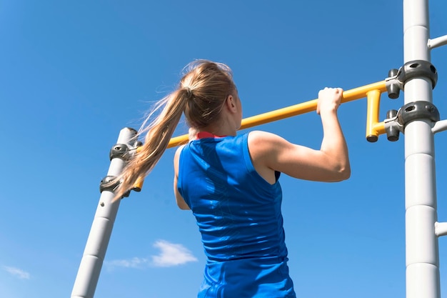 Chica haciendo ejercicio en la calle. Se sube a sí misma en la barra del campo de deportes en el parque. Foto de atrás. El hombre está irreconocible.