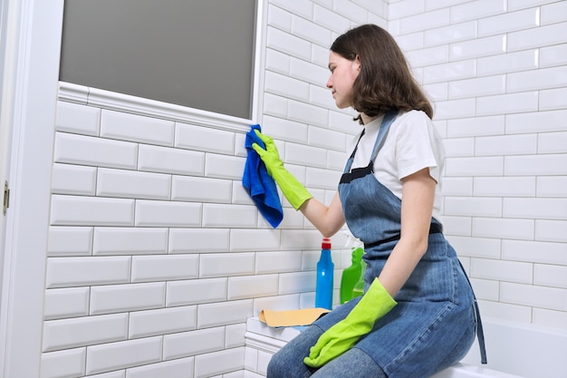 Chica haciendo limpieza en el baño. Adolescente en delantal guantes con detergente y trapo de lavar la pared de baldosas blancas, copie el espacio. Ama de llaves, limpieza en el hogar, servicio, gente joven