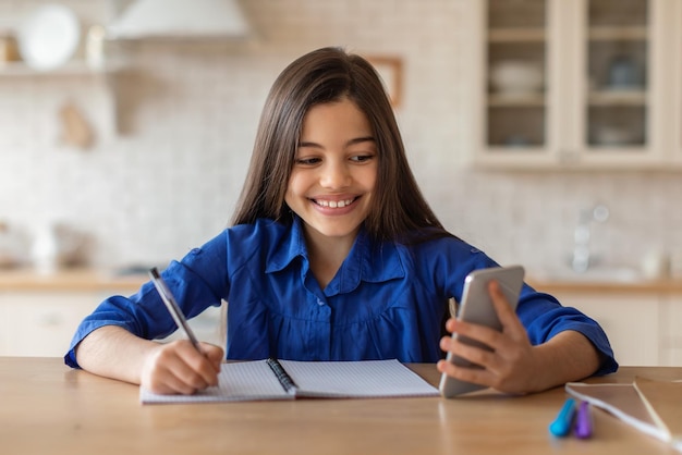 Chica haciendo tareas escolares en el teléfono inteligente tomando notas sentada en casa