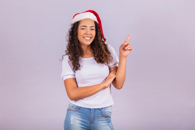 Chica hermosa y alegre con sombrero de Santa Claus apuntando a espacio libre para texto.