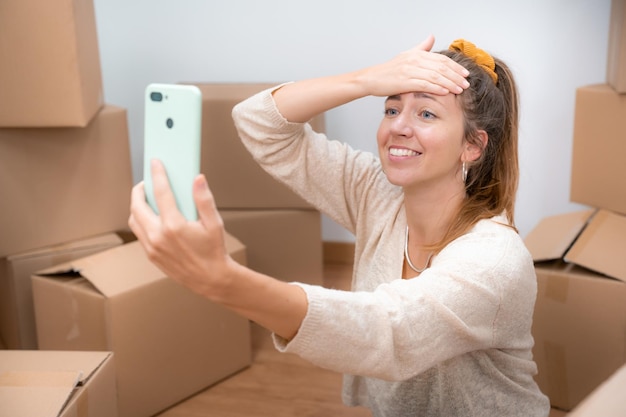 Una chica hermosa con cajas de cartón desempaquetando en su nueva casa tomando una selfie o una llamada de video