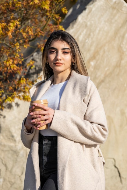 Chica hermosa confiada sosteniendo una taza de café y mirando a la cámara. Modelo de moda de mujer de retrato de belleza al aire libre