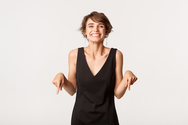 Chica hermosa eufórica mirando complacida, sonriendo y señalando con el dedo hacia el logotipo, de pie sobre blanco.