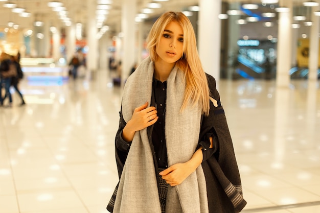 Chica hermosa joven modelo en un abrigo de moda con una bufanda caminando en el centro comercial