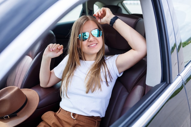 Chica hermosa de moda elegante sentada en el salón de un coche de clase ejecutiva negro. Mujer joven con una sonrisa en su rostro. Mujer en un aparcamiento