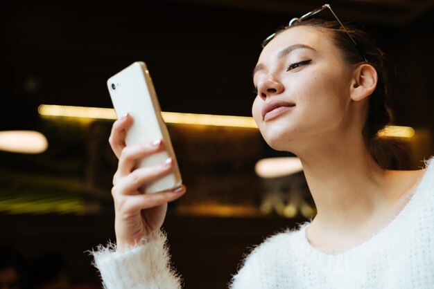 Una chica hermosa y segura mira su teléfono inteligente y sonríe sentada en un café