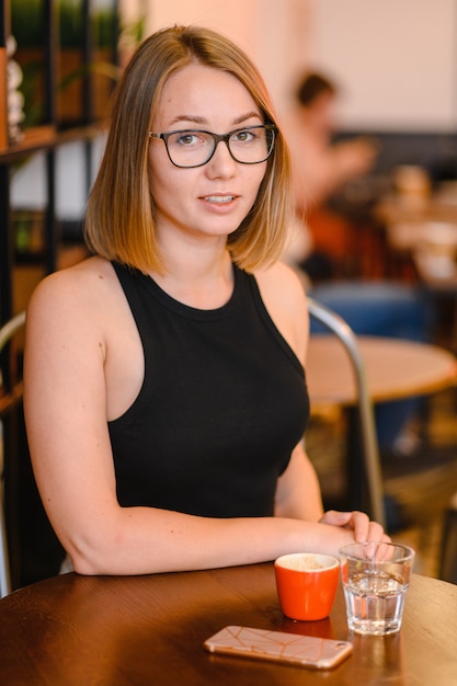 Chica con una hermosa sonrisa en gafas sentado en un café. Beber café, sosteniendo una gorra de café