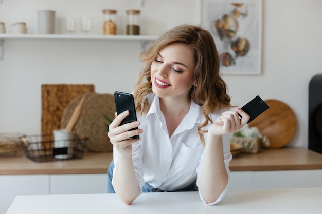 Foto una chica hermosa con un teléfono inteligente y una tarjeta bancaria en la cocina