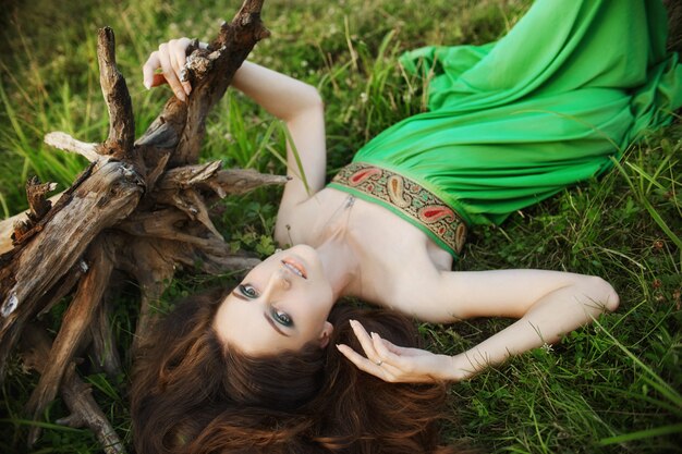 Chica en un hermoso vestido verde se encuentra en la hierba cerca de las raíces de un árbol y sueña.