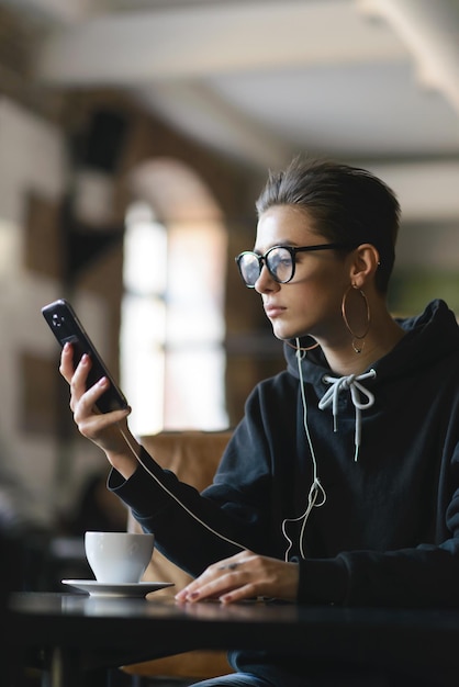 Chica hipster de atajo con gafas activa la música en su teléfono inteligente mientras se sienta en la mesa en el café