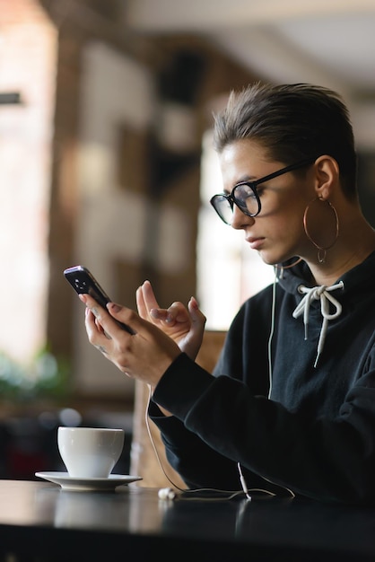 La chica hipster de atajo con gafas enciende la música en el teléfono inteligente mientras está sentada en el café