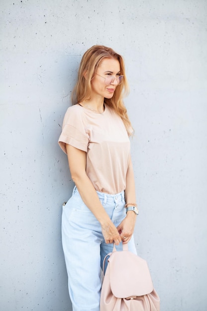 Chica hipster con camiseta beige en blanco y jeans posando contra la pared de la calle áspera
