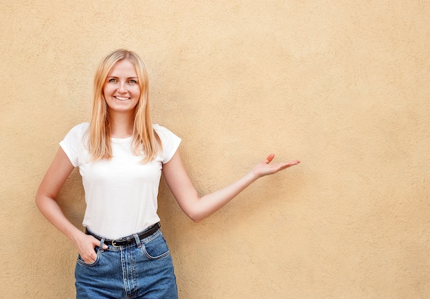 Chica hipster con camiseta blanca en blanco y jeans posando contra la pared de la calle áspera, estilo de ropa urbana minimalista, mujer muestra a mano