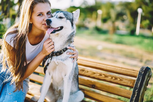 chica hipster caucásica sentada en un banco y abrazando a su adorable mascota husky en el clima soleado al aire libre