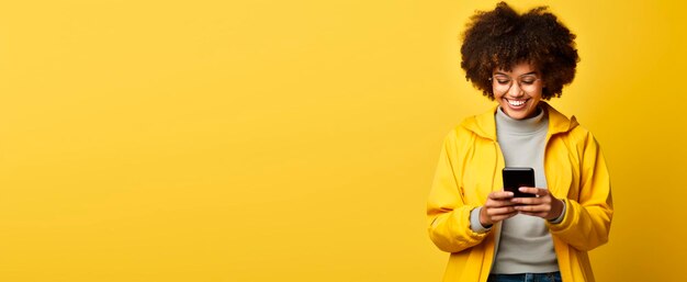 Una chica hipster con corte de pelo afro disfruta de la comunicación en línea y los servicios en línea usan chaquetas de vaqueros de moda desplazan aplicaciones de redes sociales en el dispositivo de teléfono celular aislado con espacio de copia en fondo amarillo