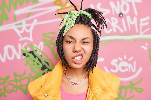 Chica hipster descarada con rastas peinadas aprieta los dientes sonríe la cara parece segura de sí misma en la cámara pasa tiempo libre en poses de la calle sobre la pared de la pared de graffiti