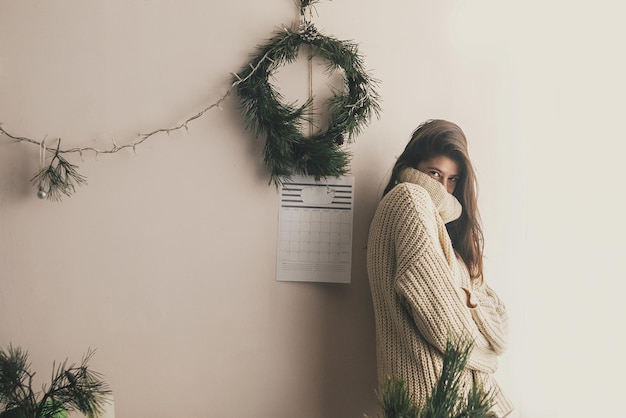 Chica hipster con estilo en suéter de punto sonriendo en una habitación rústica sobre el fondo de luces y coronas navideñas hechas a mano Momentos acogedores atmosféricos vacaciones de invierno Moody imagexA