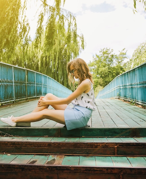 Chica hipster en falda azul y blusa blanca está sentada en el puente de madera Foto colorida de verano de una mujer de moda Estilo de vida de concepto de viaje Sesión de fotos de verano al aire libre