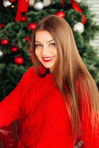 Chica hipster de invierno romántico en suéter rojo de punto cerca del árbol de Navidad