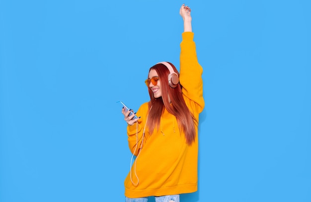 Chica hipster de moda en sudadera con capucha escuchando música y disfrutando de la vida sobre fondo azul