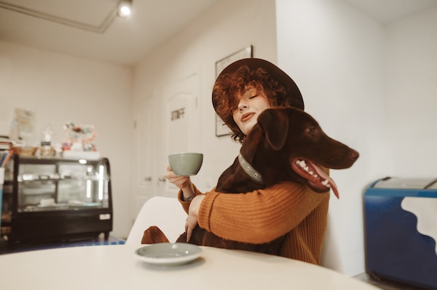 Chica hipster en ropa elegante y sombrero abraza a perro en la mesa de café y sostiene la taza de café en la mano