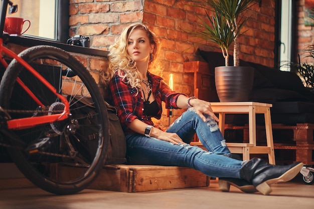 Chica hipster rubia sensual con el pelo largo y rizado vestido con una camisa de lana y jeans sentado en una caja de madera y apoyado en una pared de ladrillo, mirando hacia otro lado, en un estudio con un interior de loft.