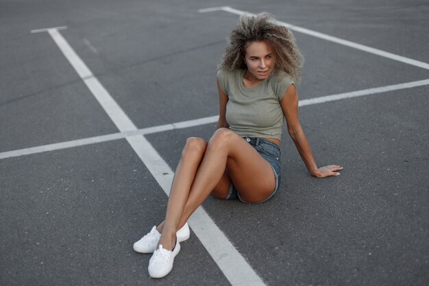 Chica hipster sexy hermosa de moda con el pelo rizado en pantalones cortos de mezclilla con una camiseta y zapatillas blancas se sienta en el asfalto en la calle