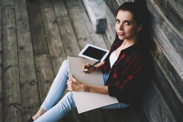 chica hipster sonriente sentada en el suelo cerca de la pared mientras escribe un ensayo con una idea emocionada en el libro de texto