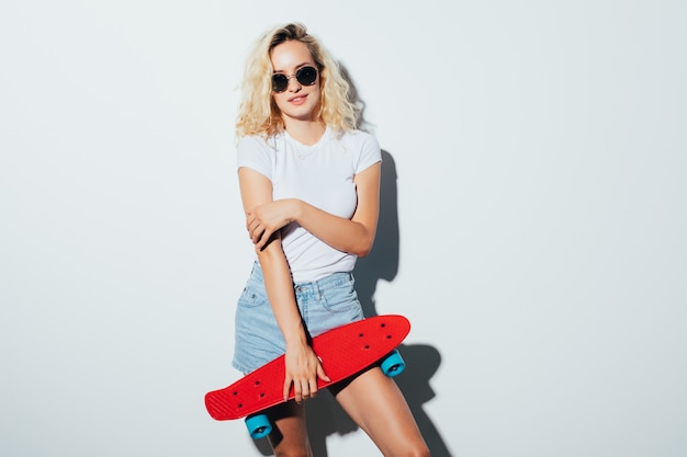 Chica hipster sosteniendo una patineta aislada en la pared blanca