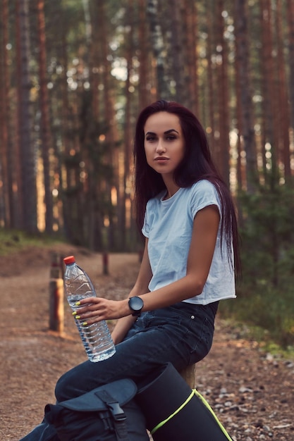 Chica hipster turista sostiene una botella de agua, se detuvo a descansar en el hermoso bosque de otoño.