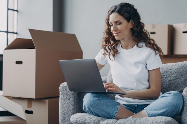 Foto chica hispana sentada con una laptop entre cajas de cartón y sonriente reubicación y trabajo en línea