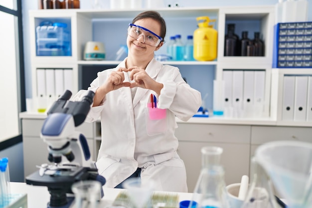 Chica hispana con síndrome de down trabajando en un laboratorio científico sonriendo enamorada haciendo forma de símbolo de corazón con las manos. concepto romántico.