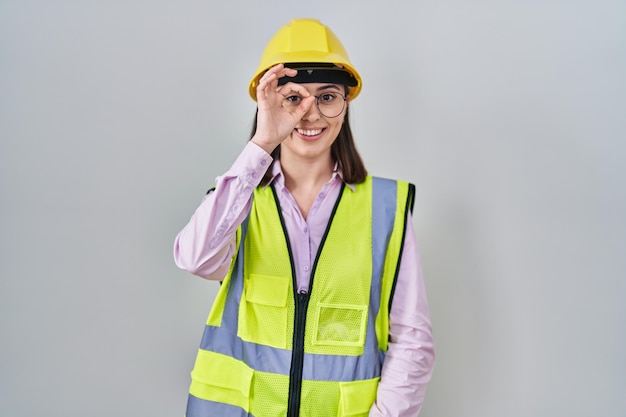 Chica hispana con uniforme de constructor y casco haciendo un buen gesto con la mano sonriendo, mirando los ojos a través de los dedos con cara feliz.