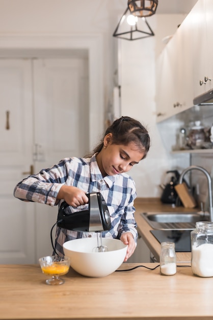 Chica horneando y mezclando masa para galletas de avena con ciruelas pasas. Niña amasando la masa con batidora o licuadora. Comida hecha en casa.