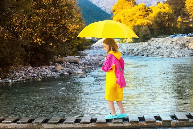 Una chica con un impermeable brillante y un paraguas amarillo cruza el río por el puente