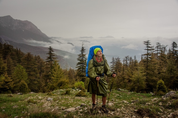 Chica en el impermeable de pie con mochila de senderismo y palos