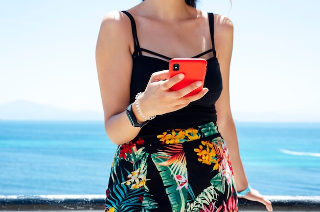 Chica inconformista mantenga en gadget de teléfono inteligente en la playa de la costa de arena, el viajero espera y usa el móvil en la mano femenina en el horizonte del paisaje marino de fondo. Mirada turística en sol azul.