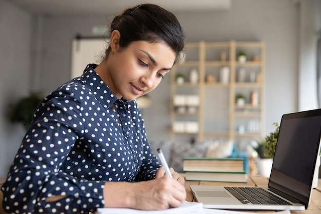 Una chica india inteligente hace notas estudiando en la computadora portátil en casa.