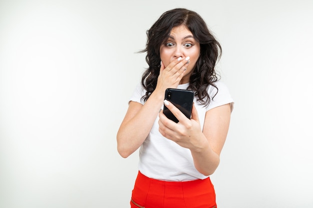 Chica inesperadamente sorprendida con una camiseta blanca con un teléfono en la mano sobre un fondo blanco de estudio.