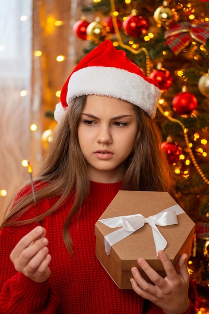 Chica infeliz con bengalas terminadas de pie cerca del árbol de Navidad con caja presente en sus manos Momentos desagradables en tiempos de felices fiestas